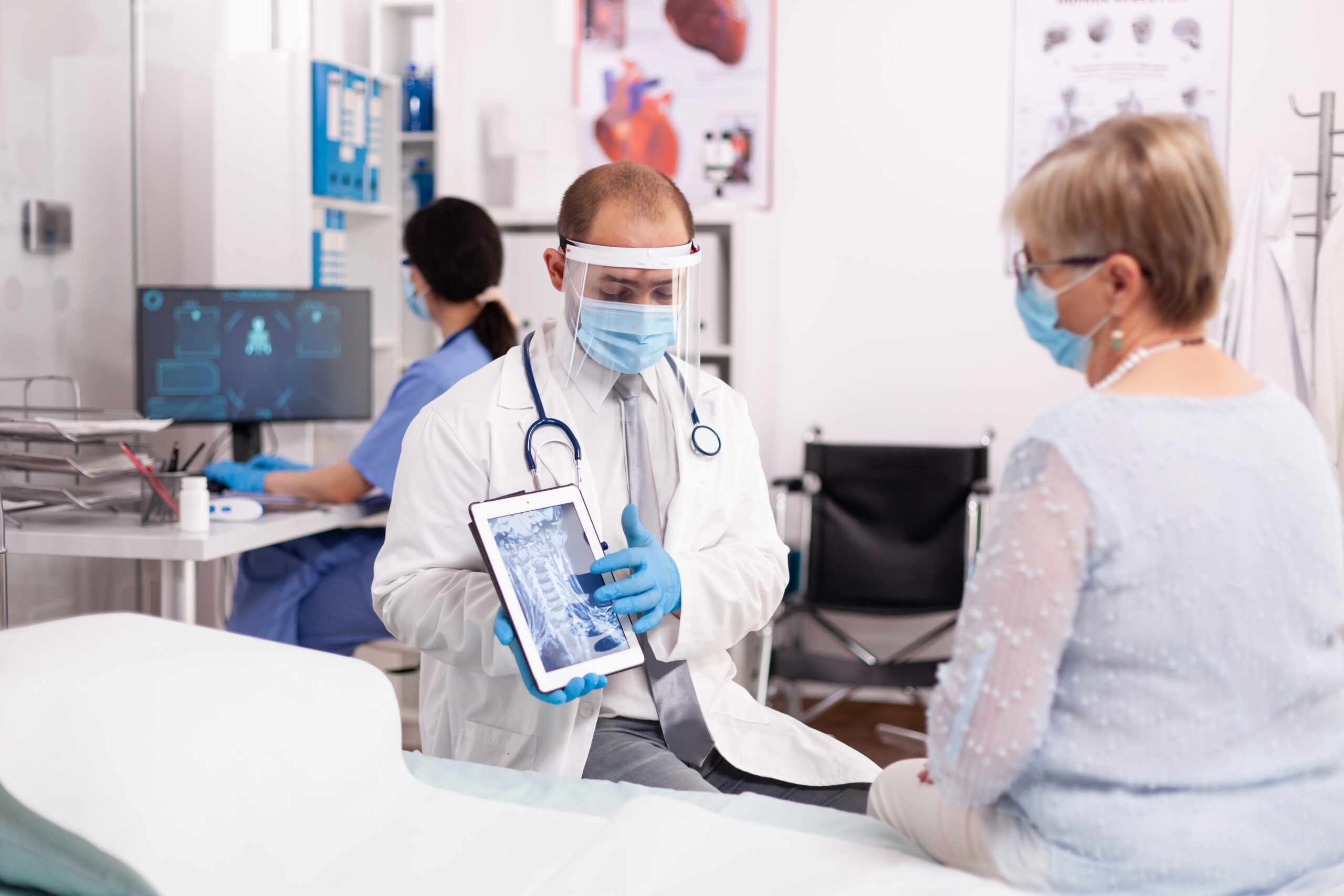 Medic explaining treatment to senior woman pointing at x-ray on tablet pc wearing face mask. Medical physician specialist during coronavirus outbreak discussing with patient.